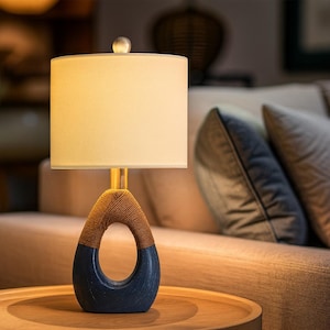 May include: A modern table lamp with a cream-colored lampshade and a unique base. The base features a dark blue, teardrop-shaped design with a woven brown rope detail. The lamp has a gold-colored stem and a small, clear orb at the top.