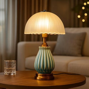 May include: A decorative table lamp with a cream-colored, ribbed lampshade and a teal and gold base. The lamp is on a wooden table, with a glass of water in the foreground. The lamp is lit, casting a warm glow.