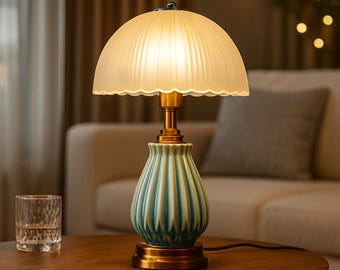 May include: A decorative table lamp with a cream-colored, ribbed lampshade and a teal and gold base. The lamp is on a wooden table, with a glass of water in the foreground. The lamp is lit, casting a warm glow.