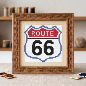 May include: A framed cross-stitch embroidery featuring the iconic Route 66 highway sign. The design displays the word ROUTE in white letters on a red background above the black number 66 on a white background, all enclosed in a blue border.