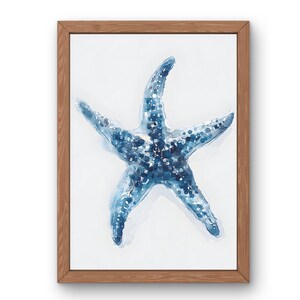 Könnte beinhalten: Ein gerahmter Aquarelldruck mit einem blauen Seestern vor weißem Hintergrund. Der Seestern hat ein strukturiertes Aussehen mit dunkleren blauen Punkten. Der Rahmen besteht aus hellbraunem Holz und verleiht dem Kunstwerk eine natürliche Note.
