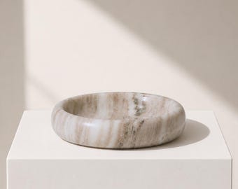 Puede incluir: Un cuenco redondo y poco profundo hecho de piedra pulida con un patrón de mármol natural que presenta vetas arremolinadas en tonos beige, crema y marrón claro. El recipiente tiene un borde redondeado liso y una estructura sólida.