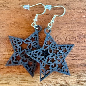 May include: A pair of black star-shaped earrings with intricate cut-out designs. The earrings have silver-colored hooks and clear plastic earring backs. The stars are made of a dark material and are displayed on a wooden surface.