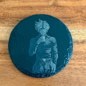 Puede incluir: Un posavasos redondo de pizarra negra con una imagen grabada de un personaje de la serie de anime Dragon Ball Z. El personaje se representa en una pose dinámica, sosteniendo un objeto pequeño. El posavasos tiene aproximadamente 10 cm de diámetro.