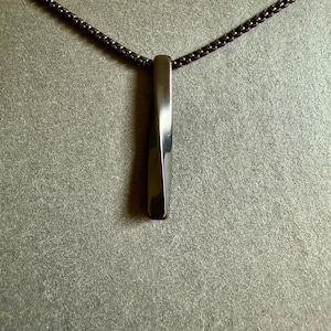 May include: A silver-toned pendant necklace featuring a rectangular, slightly twisted pendant. The pendant hangs from a dark, chain-style necklace. The jewelry is displayed against a neutral gray background.