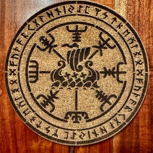 May include: Circular coaster with a brown base and a black Viking symbol design. The design includes a ship, runes, and other symbols within a circular border. The coaster is made of a cork-like material.