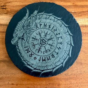 May include: Round, black slate coaster featuring a silver dragon design encircling a central rune symbol. The dragon's head is visible on the left, with intricate details. The coaster has a textured surface and is suitable for beverages.