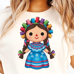 May include: A graphic of a traditional Mexican doll, wearing a blue dress with white floral details and a colorful headband. The doll has brown braids and a smiling face. The design is printed on a cream-colored t-shirt.
