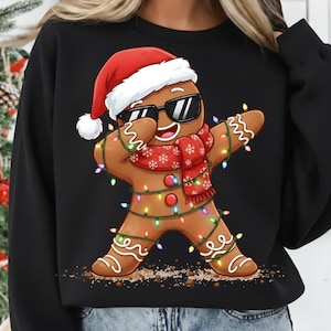 May include: Black sweatshirt featuring a gingerbread man wearing sunglasses, a Santa hat, and a red scarf with snowflakes. The gingerbread man is decorated with colorful Christmas lights and is in a dabbing pose.
