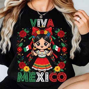 May include: Black sweatshirt featuring a cartoon girl in a traditional Mexican dress, holding Mexican flags. The design includes the words "VIVA MEXICO" in colorful lettering, surrounded by roses and floral accents. The girl has black braids and is wearing a flower crown.