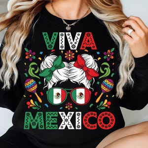 Op de afbeelding: Zwart sweatshirt met de tekst "VIVA MEXICO" in wit, groen en rood. Het ontwerp bevat een vrouwengezicht met groene en rode strikken, een zonnebril met de Mexicaanse vlag en maracas. Kleurrijke bloemen en decoratieve elementen omringen de centrale afbeelding.