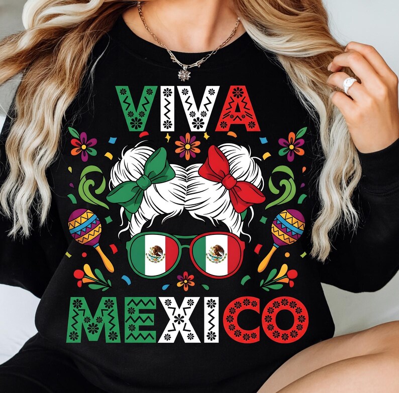 Op de afbeelding: Zwart sweatshirt met de tekst "VIVA MEXICO" in wit, groen en rood. Het ontwerp bevat een vrouwengezicht met groene en rode strikken, een zonnebril met de Mexicaanse vlag en maracas. Kleurrijke bloemen en decoratieve elementen omringen de centrale afbeelding.