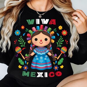 Op de afbeelding: Zwart sweatshirt met de woorden "VIVA MEXICO" in kleurrijke letters. Het ontwerp toont een cartoonpop in een blauwe jurk, vlechten en een kleurrijke hoofdband, met maracas, omringd door bloemen.