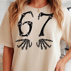 May include: Beige t-shirt with a black skeleton design. The design features the number "67" in a bone-like font, with two skeleton hands below. The shirt is made of a soft, comfortable material.