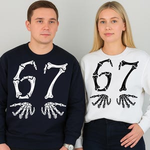Puede incluir: Dos sudaderas de cuello redondo, una azul marino y otra blanca. Cada sudadera presenta el número "67" en una fuente de hueso, con manos esqueléticas debajo. Las sudaderas son usadas por dos personas.