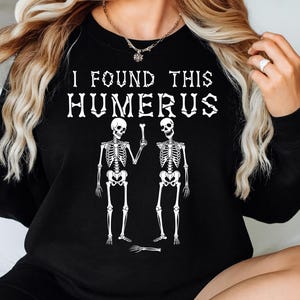 May include: Black sweatshirt with the text "I FOUND THIS HUMERUS" in white, featuring two skeleton figures. One skeleton holds a bone, and a detached hand is at the bottom. A fun, spooky design.