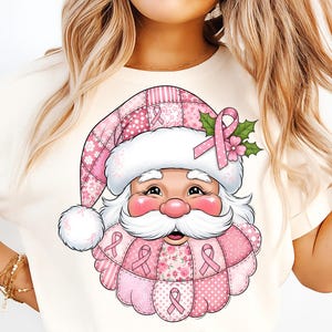 May include: A cream-colored t-shirt featuring a Santa Claus graphic with a pink ribbon theme. Santa's hat and beard are adorned with pink ribbons and floral patterns. The design includes a pink ribbon and holly leaves.