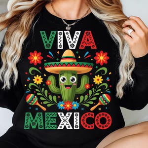 Op de afbeelding: Zwart sweatshirt met een kleurrijk "VIVA MEXICO" ontwerp. De afbeelding toont een lachende cactus met een sombrero, omringd door bloemen en maracas. Het ontwerp gebruikt groen, rood, geel en blauw.