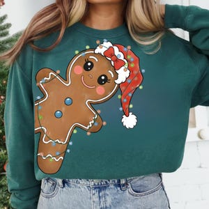 Pode incluir: Camisola verde-azulado com um desenho de homem de gengibre. O homem de gengibre usa um gorro de Pai Natal e é decorado com luzes coloridas. Um design festivo, ideal para a época natalícia.
