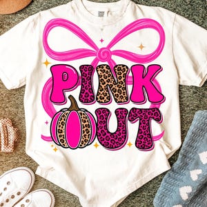 Könnte beinhalten: Weißes T-Shirt mit einer rosa Schleife und dem Schriftzug "PINK OUT" in fetter, verspielter Schrift. Die Buchstaben "PINK" haben ein Leopardenmuster. Ein rosa Kürbis ist ebenfalls abgebildet.