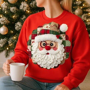 Könnte beinhalten: Roter Weihnachts-Sweatshirt mit einem Santa Claus-Applikation. Das Santa-Design hat einen weißen Bart, runde Brille und eine rote Nase. Die Applikation ist von einem Patchwork aus karierten und floralen Stoffen umgeben. Eine weiße Tasse wird gehalten.