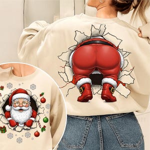 May include: Beige sweatshirt with a graphic of Santa Claus breaking through a wall. The front shows Santa's face, while the back features his red pants and boots. Festive Christmas apparel.