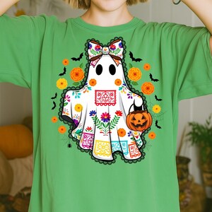 May include: Green t-shirt featuring a white ghost design with a floral bow, holding a jack-o'-lantern. The ghost is decorated with colorful flowers and Day of the Dead-inspired patterns. Orange flowers and black bats surround the ghost.