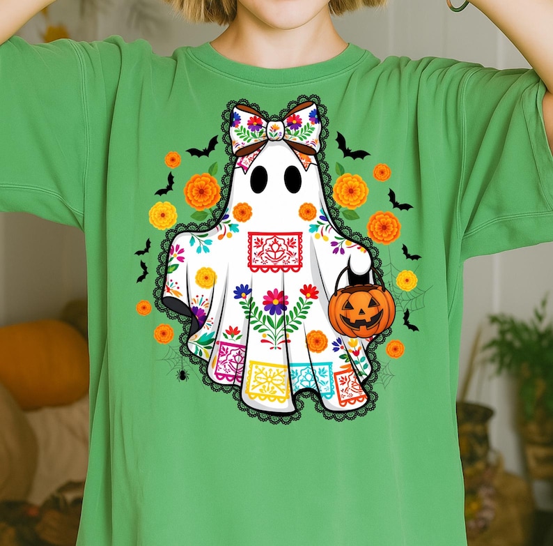 May include: Green t-shirt featuring a white ghost design with a floral bow, holding a jack-o'-lantern. The ghost is decorated with colorful flowers and Day of the Dead-inspired patterns. Orange flowers and black bats surround the ghost.