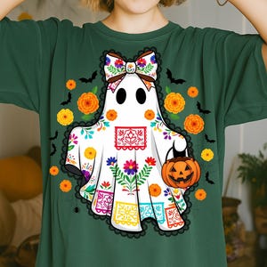 May include: A white ghost design on a dark green t-shirt. The ghost is decorated with colorful floral patterns, a bow, and a jack-o'-lantern. Black bats and orange flowers surround the ghost, creating a festive Halloween theme.