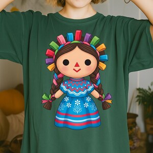 K&ouml;nnte beinhalten: Gr&uuml;nes T-Shirt mit einer farbenfrohen Illustration einer traditionellen mexikanischen Puppe. Die Puppe hat ein gro&szlig;es Gesicht, geflochtene Haare und ein blaues Kleid mit floralen Details. Sie tr&auml;gt ein dekoratives Stirnband mit bunten B&auml;ndern.
