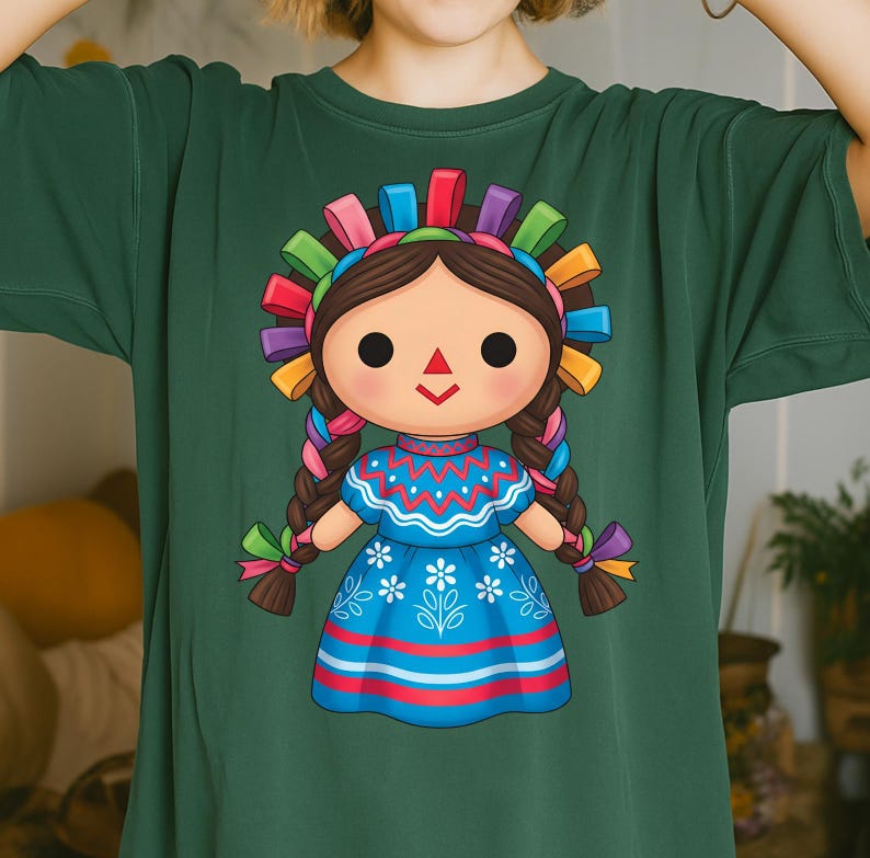 K&ouml;nnte beinhalten: Gr&uuml;nes T-Shirt mit einer farbenfrohen Illustration einer traditionellen mexikanischen Puppe. Die Puppe hat ein gro&szlig;es Gesicht, geflochtene Haare und ein blaues Kleid mit floralen Details. Sie tr&auml;gt ein dekoratives Stirnband mit bunten B&auml;ndern.
