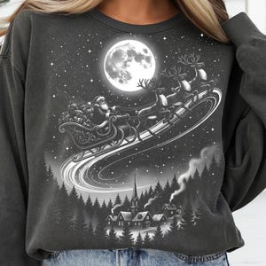 May include: Dark gray sweatshirt featuring a black and white illustration of Santa Claus in his sleigh, pulled by reindeer, flying across a starry night sky with a full moon. A village is nestled below.