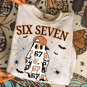 May include: A light beige sweatshirt with the words "SIX SEVEN" in black and orange. The graphic features a ghost wearing an orange baseball cap and holding up two fingers. Spiders, spiderwebs, and bats are also featured.