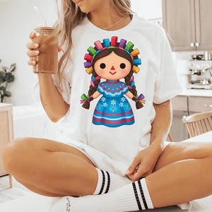 K&ouml;nnte beinhalten: Wei&szlig;es T-Shirt mit einem Cartoon-Puppe-Design. Die Puppe hat braune Z&ouml;pfe, ein blaues Kleid mit floralen Details und einen bunten Kopfschmuck. Das Shirt wird mit wei&szlig;en Socken mit schwarzen Streifen getragen.