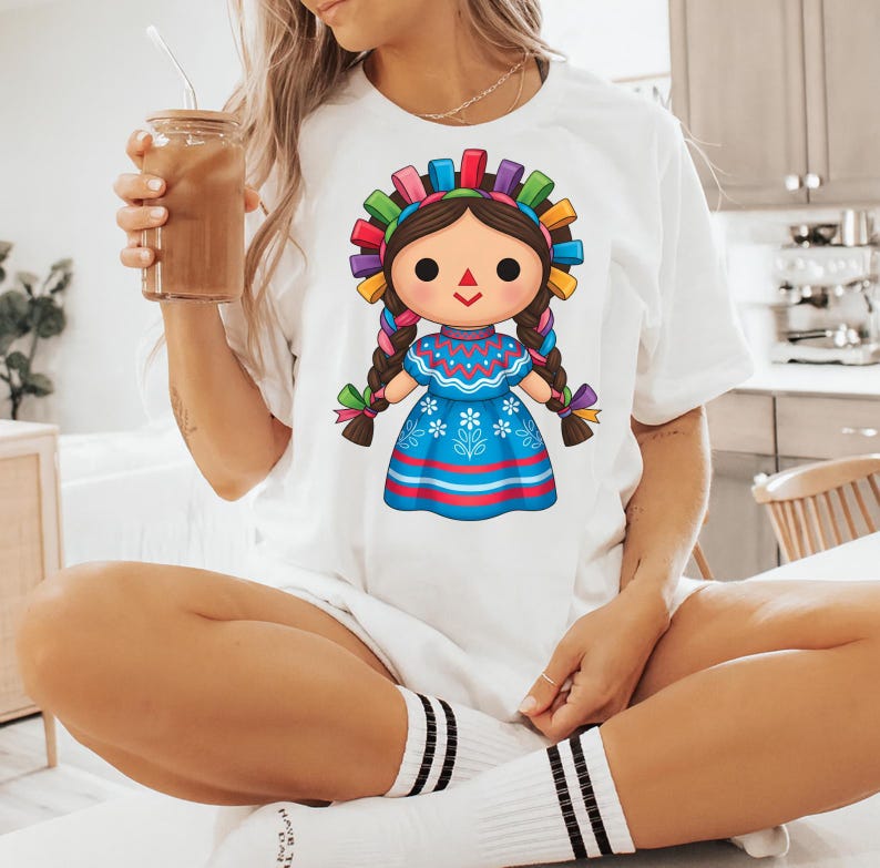 K&ouml;nnte beinhalten: Wei&szlig;es T-Shirt mit einem Cartoon-Puppe-Design. Die Puppe hat braune Z&ouml;pfe, ein blaues Kleid mit floralen Details und einen bunten Kopfschmuck. Das Shirt wird mit wei&szlig;en Socken mit schwarzen Streifen getragen.