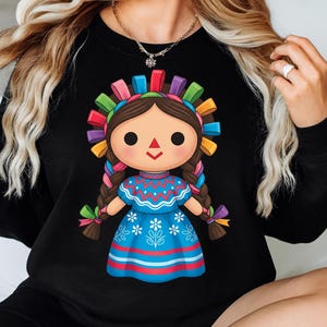 K&ouml;nnte beinhalten: Schwarzes Sweatshirt mit einer farbenfrohen Illustration einer traditionellen mexikanischen Puppe. Die Puppe hat braune Z&ouml;pfe, ein blaues Kleid mit wei&szlig;en floralen Details und einen lebendigen Kopfschmuck mit Regenbogenb&auml;ndern. Das Design ist mittig platziert.