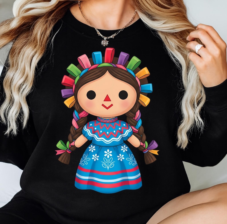 K&ouml;nnte beinhalten: Schwarzes Sweatshirt mit einer farbenfrohen Illustration einer traditionellen mexikanischen Puppe. Die Puppe hat braune Z&ouml;pfe, ein blaues Kleid mit wei&szlig;en floralen Details und einen lebendigen Kopfschmuck mit Regenbogenb&auml;ndern. Das Design ist mittig platziert.