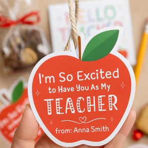 Può includere: Etichetta regalo a forma di mela rossa con la scritta "I'm So Excited to Have You As My TEACHER" e "from: Anna Smith". L'etichetta ha una foglia verde e uno spago per appenderla. Sullo sfondo una matita e un sacchetto regalo.