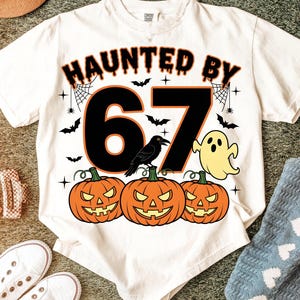 May include: White t-shirt with a Halloween design. The shirt displays the text "HAUNTED BY 67" in orange and black, accompanied by jack-o'-lanterns, bats, a ghost, and a crow.