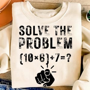 May include: A cream-colored sweatshirt with the text "SOLVE THE PROBLEM" in distressed black lettering. Below the text is the math problem (10*6)+7=?, with a pointing finger graphic. The sweatshirt is a casual style.