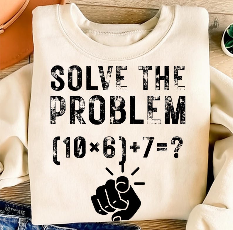 May include: A cream-colored sweatshirt with the text "SOLVE THE PROBLEM" in distressed black lettering. Below the text is the math problem (10*6)+7=?, with a pointing finger graphic. The sweatshirt is a casual style.