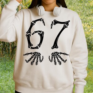 May include: Cream-colored sweatshirt featuring a black skeleton bone design. The design includes the numbers "67" in a bone font, along with two skeletal hands. The sweatshirt has a crew neck and long sleeves, perfect for Halloween or spooky-themed events.