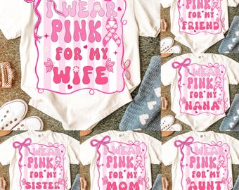 May include: Cream-colored t-shirts and onesies with pink text and graphics. The shirts say "I Wear Pink For My Wife", "I Wear Pink For My Friend", "I Wear Pink For My Nana", "I Wear Pink For My Sister", "I Wear Pink For My Mom", and "I Wear Pink For My Aunt".