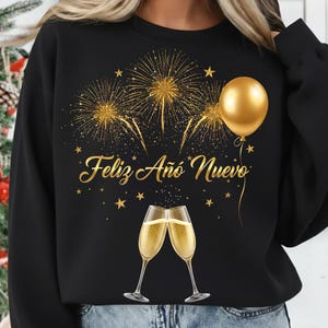 May include: Black sweatshirt with a gold design featuring fireworks, a balloon, stars, and two champagne flutes. The text "Feliz Año Nuevo" is written in gold script. A festive design for New Year's celebrations.