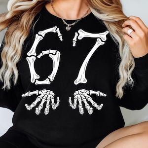 May include: Black sweatshirt with a white skeleton design. The design features the number 67 in a bone-like font, with two skeleton hands at the bottom. The sweatshirt is a crew neck style.