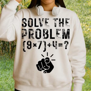 May include: Cream-colored sweatshirt with the text "SOLVE THE PROBLEM" above a math equation and a pointing finger graphic. The equation reads (9*7)+4=? The sweatshirt has a crew neck and long sleeves.