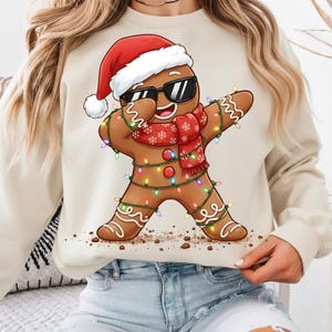 May include: A beige sweatshirt featuring a gingerbread man dabbing. The gingerbread man wears a Santa hat, sunglasses, and a red scarf with snowflakes. He is decorated with colorful Christmas lights.
