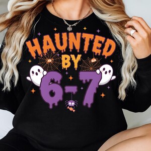 Może przedstawiać: Czarna bluza z motywem Halloween z napisem "HAUNTED BY 6-7" w kolorze pomarańczowym i fioletowym. Projekt zawiera pajęczyny, duchy i pająka. Bluza ma okrągły dekolt.