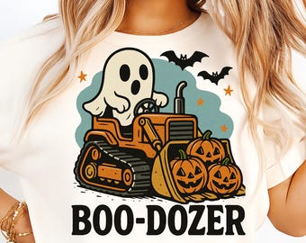 May include: Cream-colored t-shirt featuring a Halloween-themed graphic. The design includes a ghost driving an orange bulldozer with pumpkins in the front. The text "BOO-DOZER" is printed below the image, with bats and stars.