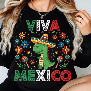 Op de afbeelding: Zwart sweatshirt met een kleurrijke afbeelding van een groene dinosaurus met een sombrero. De tekst "VIVA MEXICO" is weergegeven in wit en met kleurrijke bloemmotieven. Het ontwerp bevat maracas en bloemen.